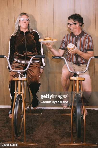 retro nineteen eighties aesthetic exercise bike couple with man eating donuts - funky middle age couple stockfoto's en -beelden