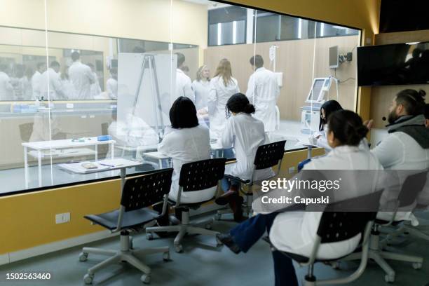 medizinstudenten beobachten den praktischen unterricht von außerhalb des klassenzimmers durch ein glasfenster - medizinische fakultät stock-fotos und bilder