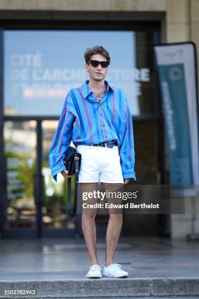 Guest wears black sunglasses, a flashy blue and red striped print pattern shirt, a black shiny leather belt, a black shiny leather handbag, white...