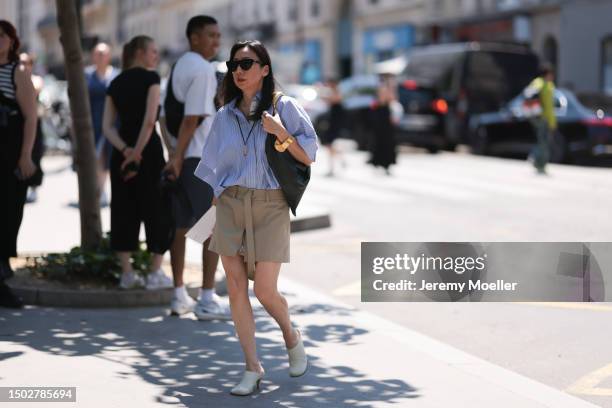 Fashion Week Guest is seen wearing black shades, a blue white and black striped shirt, a black bag with golden ornaments, a beige shorts with a long...