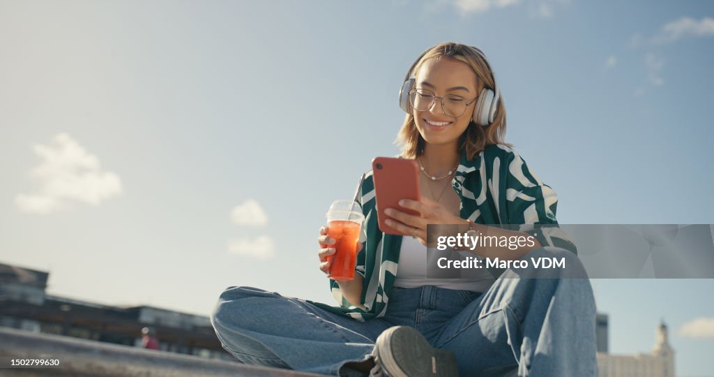 Young girl, headphones and cellphone in city with cool drink, mobile texting and social media app on mockup sky. Happy gen z woman, urban influencer and listening to music with smartphone outdoor