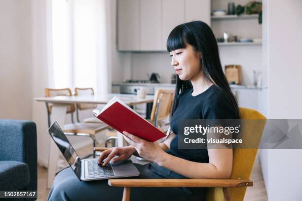 female asian woman reading book and making notes at home, concentrating on her research. - reference book stock pictures, royalty-free photos & images