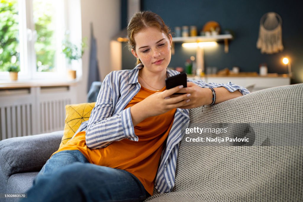 Rothaariges kaukasisches Teenager-Mädchen mit Handy, während es auf dem Sofa sitzt