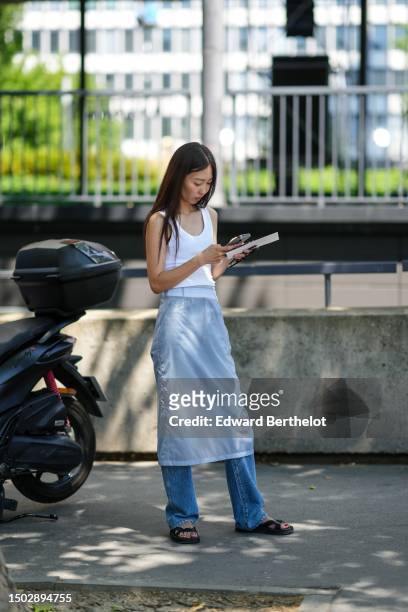 Guest wears a white tank-top, a pale blue midi tulle skirt, blue denim flared pants, black shiny leather sandals , outside Sacai, during the Menswear...