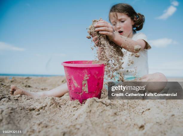 a little girls scoops handfuls of sand from the beach and drops it into her plastic pink pail - bucket and spade stock pictures, royalty-free photos & images