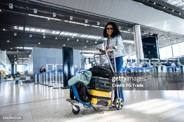 retrato de una mujer adulta mediana usando el teléfono mientras camina con un carrito de equipaje en el aeropuerto - carrito-para-equipaje fotografías e imágenes de stock