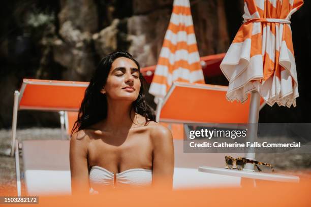 a serene woman enjoys the feeling of sun on her face, as she sits on a sun lounger on a beach - tomar el sol fotografías e imágenes de stock