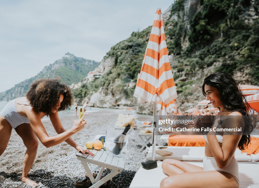 A woman takes a photo of her female friend on a sun lounger on a beach, using her phone