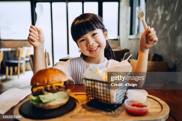 cheerful little asian girl smiling joyfully at the camera while enjoying eating burger with fries in a restaurant. can't wait digging in. eating out lifestyle - child foodie photos et images de collection