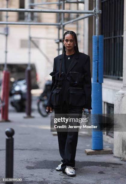 Guest is seen wearing black sunglasses, a black shiny suit with big attached bags and zippers on the blazer combined with black Botter shirt with...