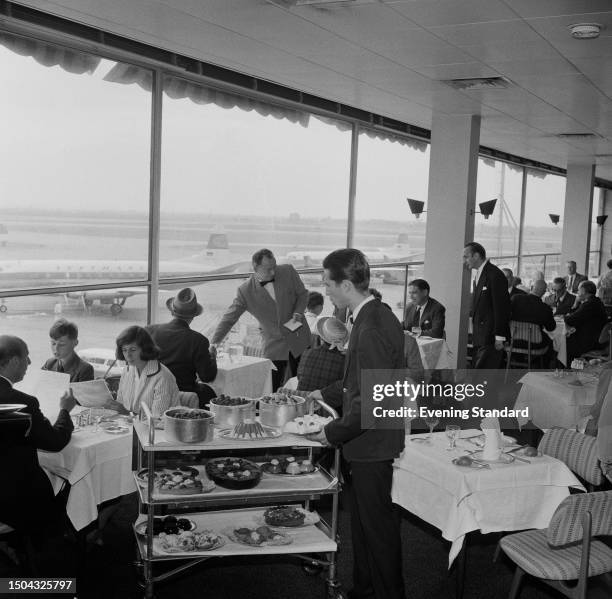 Waiter approaches diners with a food trolley of desserts at London Airport's Forte's restaurant, with windows overlooking airplanes, August 11th 1959.