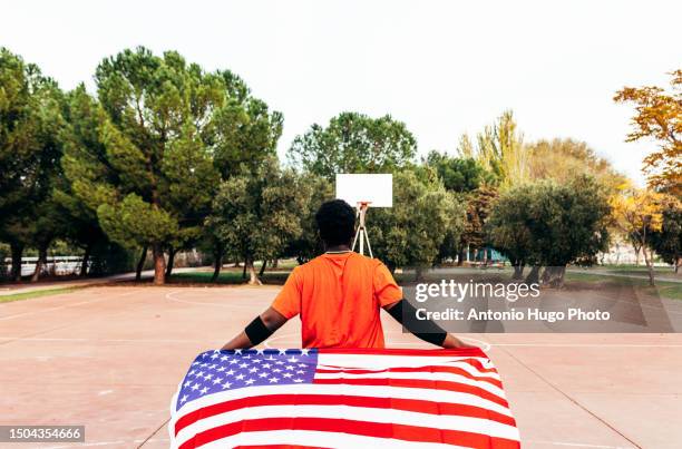 black african-american boy carrying the u.s. flag on an urban basketball court. dressed with an orange t-shirt. - u know stock pictures, royalty-free photos & images