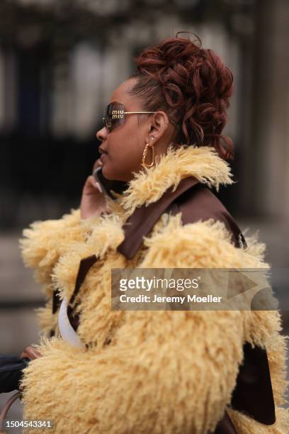 Fashion Week guest is wearing a golden Miu Miu Sunglasses with brown lenses, a pair of golden earrings/hoops and many ear studs, a brown jacket with...