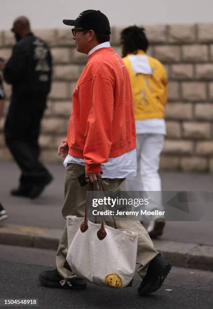 Fashion Week guest is wearing black sunglasses, a black cap, a white tee with an orange sweater below it, khaki wide pants, black sneakers and a tote...