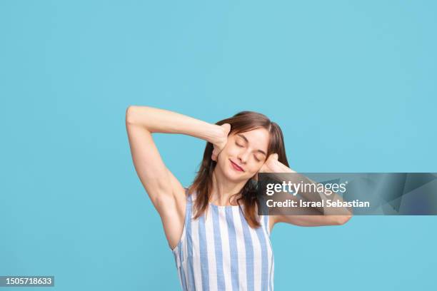 conceptual portrait of a young caucasian woman in a striped jumpsuit covering her ears with her eyes closed against a blue wall. - woman hush stock pictures, royalty-free photos & images