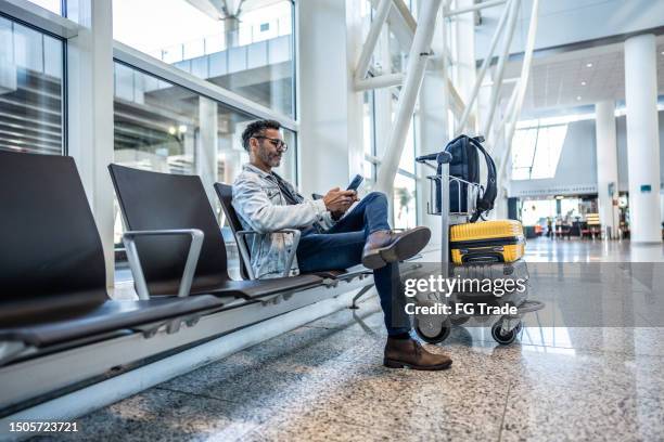 hombre maduro usando teléfono móvil en la sala de espera del aeropuerto - carrito-para-equipaje fotografías e imágenes de stock
