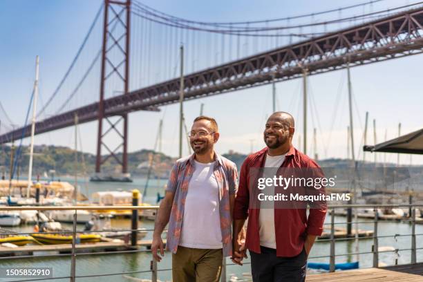 gay couple walking hand in hand in front of the 25th of april bridge - casal gay imagens e fotografias de stock