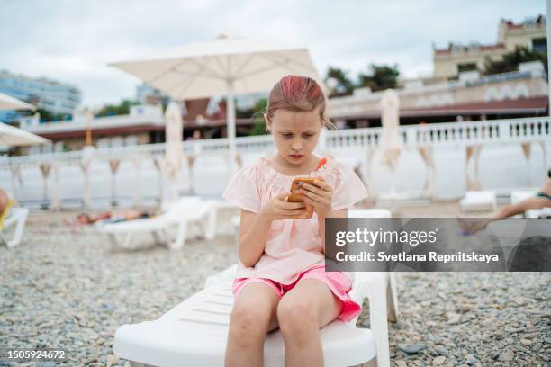 child using smartphone sitting in sun lounger on beach sea - smartphone strand stock-fotos und bilder