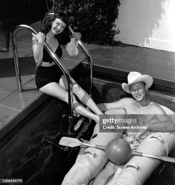 Roy Rogers wearing a stetson as he relaxes in an inflatable with Dale Evans posing beside him in a bikini at their home swimming pool, circa 1950.