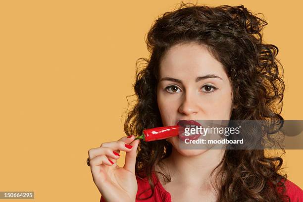 portrait of a young woman biting red chili pepper over colored background - chili woman fotografías e imágenes de stock