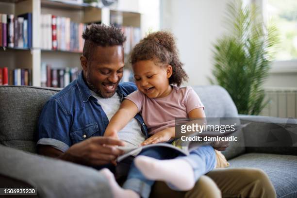 felice padre afroamericano e adorabile figlia di razza mista stanno leggendo un libro e sorridendo mentre trascorrono del tempo insieme a casa. concetto di educazione e sviluppo dei bambini - mixed magazine foto e immagini stock