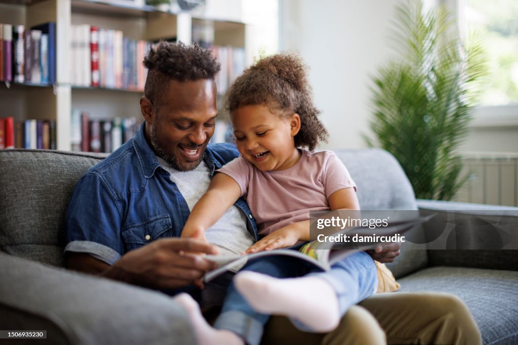 Happy African American father and adorable mixed race daughter are reading a book and smiling while spending time together at home. Children education and development concept