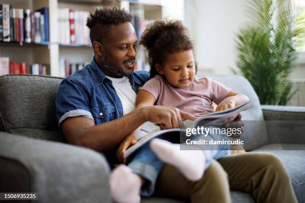 ein glücklicher afroamerikanischer vater und eine entzückende gemischtrassige tochter lesen ein buch und lächeln, während sie zu hause zeit miteinander verbringen. erziehungs- und entwicklungskonzept für kinder - mixed magazine stock-fotos und bilder