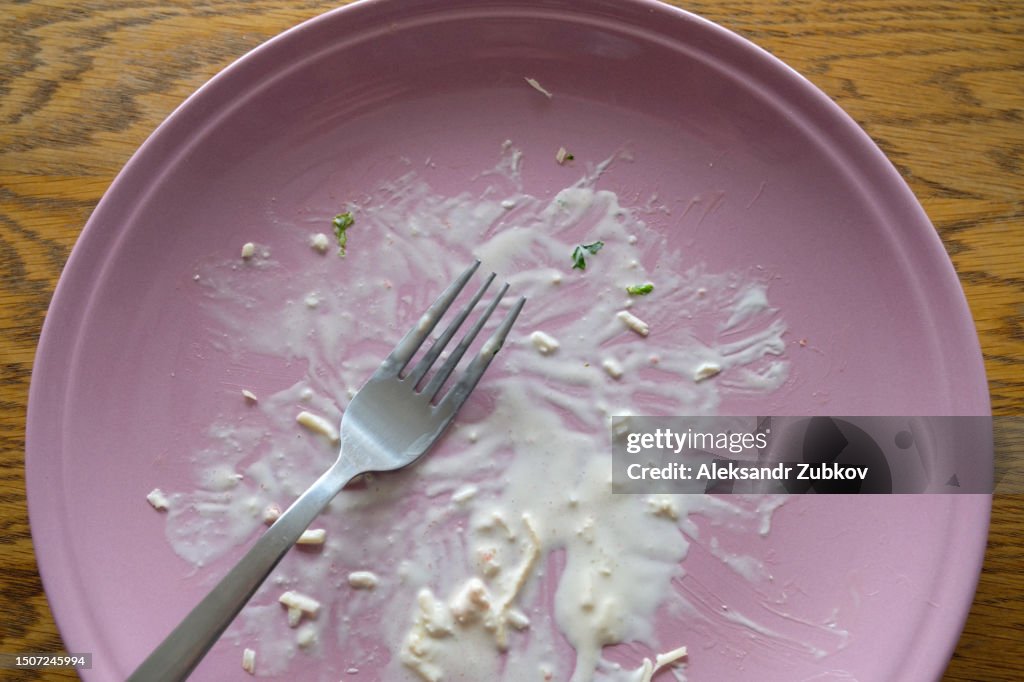 A dirty empty plate and fork on a wooden kitchen or dining table. Cutlery is used, symbolizing the end of lunch or dinner. A portion of food eaten, overeating.