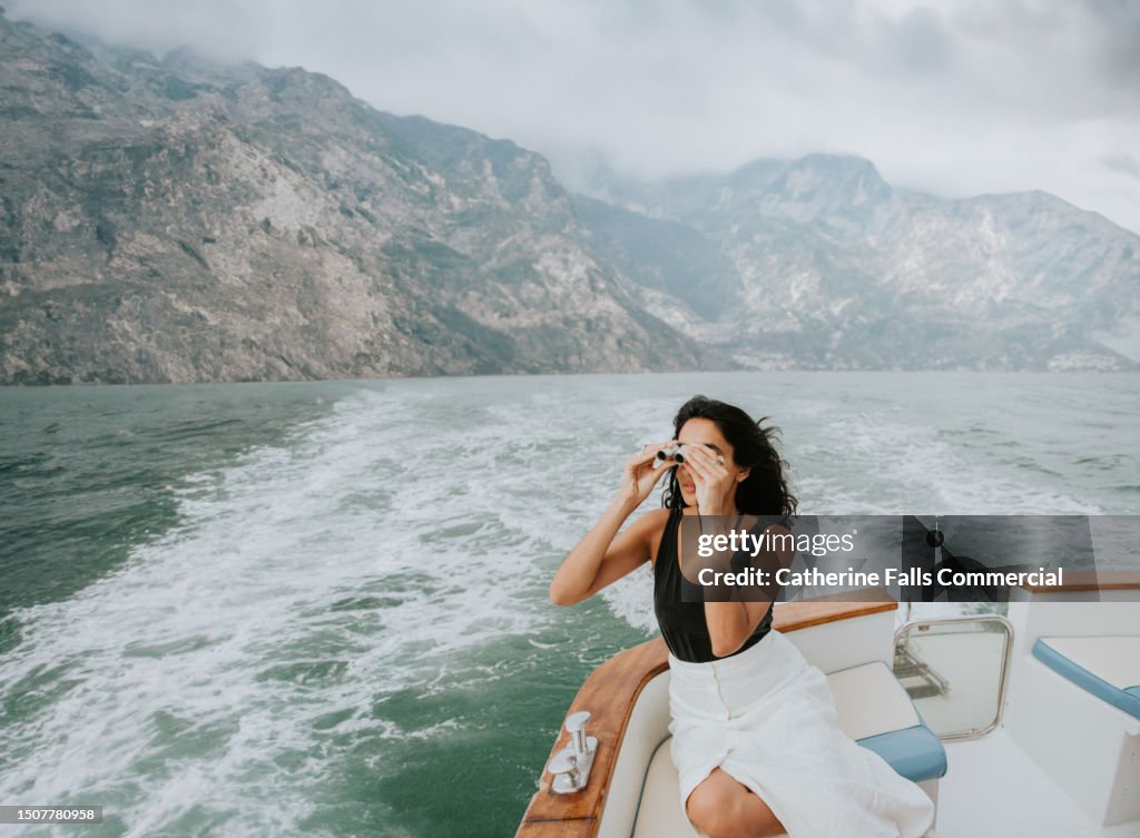 A beautiful Indian woman reclines on the stern of a small luxury yacht, and looks through a pair of small binoculars, and gazes out to sea.