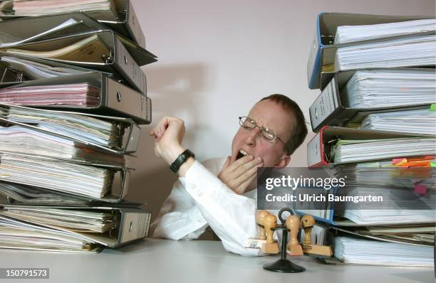 Man sitting between piles of files looks yawning at the watch, Symbolic photo for the topics: no involvement of the employees, bureaucracy, overtime.