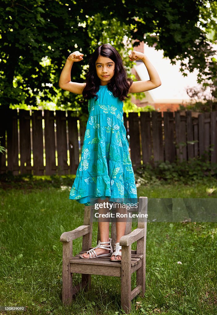 Young girl flexing muscles while standing on chair