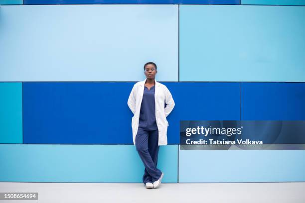 a young female doctor or medical student, donning medical attire and a white coat, leaning confidently against a blue wall, embodying the professionalism and knowledge associated with the medical field. - medizinstudent stock-fotos und bilder