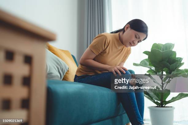 asian woman suffering from knee pain sitting on sofa in living room at home. - beautiful asian legs stockfoto's en -beelden