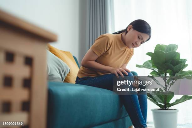 mulher asiática que sofre de dor no joelho sentada no sofá na sala de estar em casa. - beautiful asian legs - fotografias e filmes do acervo