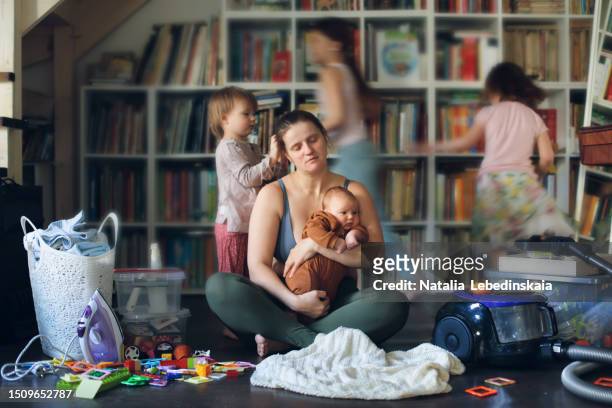 real-life parenting: fatigued mother finding solace on living room floor amidst running kids and household clutter. mom with four kids with closed eyes - madre ama de casa fotografías e imágenes de stock
