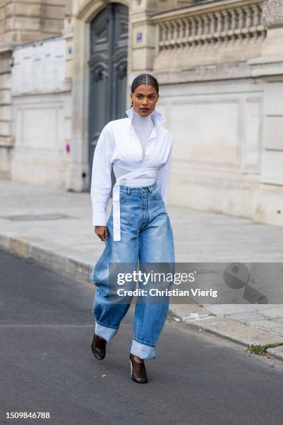 Tina Kunakey wears white wrapped blouse, turtleneck, wide leg cuffed denim jeans, heels outside Alaia on July 02, 2023 in Paris, France.