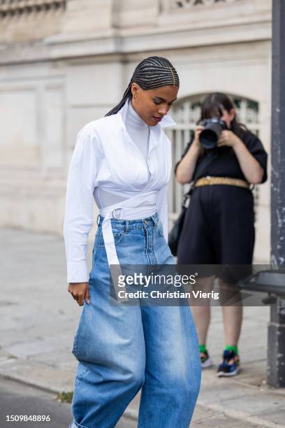 Tina Kunakey wears white wrapped blouse, turtleneck, wide leg cuffed denim jeans, heels outside Alaia on July 02, 2023 in Paris, France.