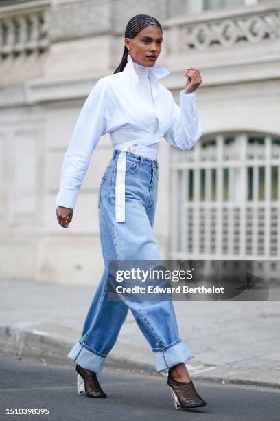 Tina Kunakey wears gold earrings, a white turtleneck long sleeves t-shirt, a white laces waist cropped waist shirt, high waist blue faded denim puffy...