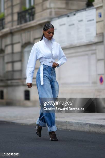 Tina Kunakey wears gold earrings, a white turtleneck long sleeves t-shirt, a white laces waist cropped waist shirt, high waist blue faded denim puffy...