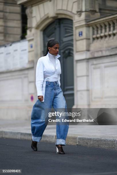 Tina Kunakey wears gold earrings, a white turtleneck long sleeves t-shirt, a white laces waist cropped waist shirt, high waist blue faded denim puffy...