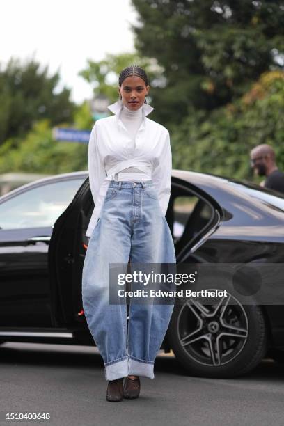 Tina Kunakey wears gold earrings, a white turtleneck long sleeves t-shirt, a white laces waist cropped waist shirt, high waist blue faded denim puffy...