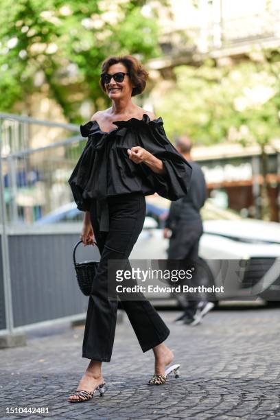 Ines de la Fressange wears black sunglasses, a black ruffled shoulder-off / puffy long sleeves silk blouse, black denim flared pants, a black braided...