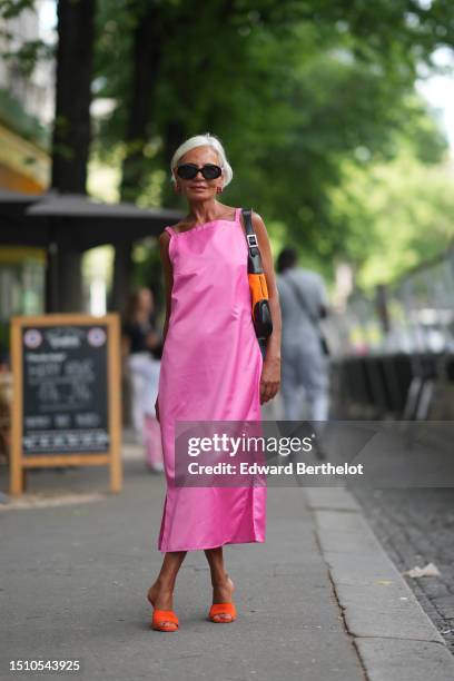 Grece Ghanem wears black sunglasses, a neon pink silk sleeveless long dress, a neon orange and black shiny leather shoulder bag, neon orange shiny...