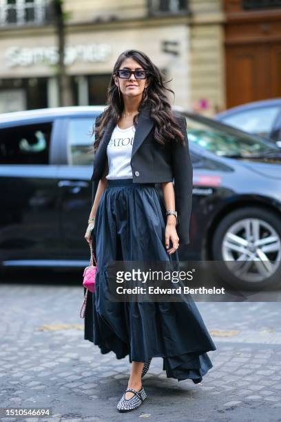 Guest wears black sunglasses, gold large pendant earrings, a white with black logo print pattern t-shirt from Patou, a black cropped jacket, a black...
