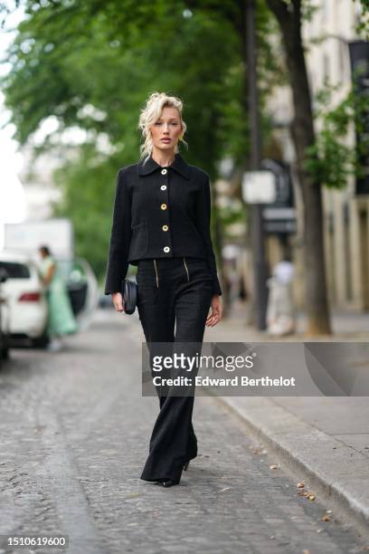 Guest wears gold earrings, a black tweed with large gold buttons jacket, a black zipper waist flared pants, a black shiny leather handbag, black...