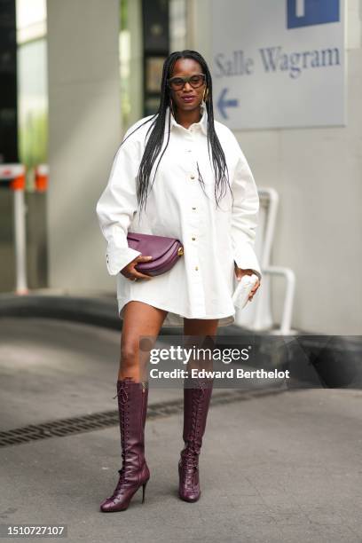 Guest wears khaki sunglasses, a white oversized short shirt dress, a burgundy shiny leather handbag from Patou, matching burgundy shiny leather heels...