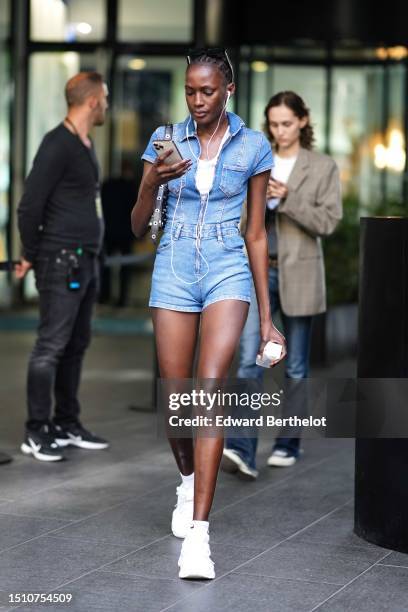 Guest wears black glasses, a white tank-top, a pale blue denim short sleeves belted short jumpsuit, a black shiny leather nailed / studded shoulder...