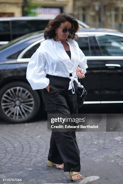 Guest wears black circle sunglasses, a black shoulder-off top, a white shirt, black large cargo pants, a black cut-out puffy crossbody bag, gold...