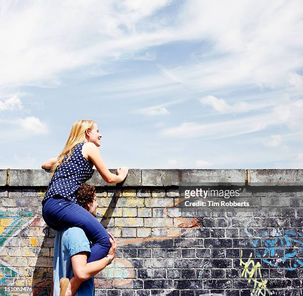 man helping young woman look over wall. - neugierde stock-fotos und bilder