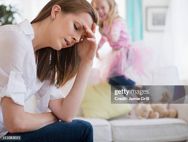 mother with headache, daughter in background - stressed young woman sitting on couch stock-fotos und bilder